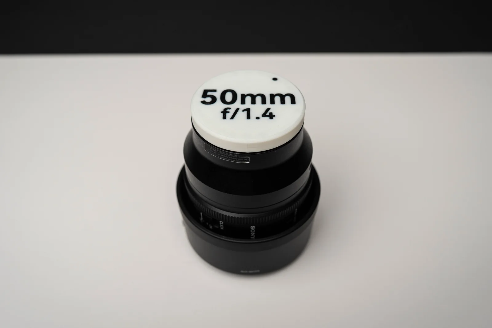 A white 3D-printed rear lens cap marked 50mm f/1.4 in black, fitted to a black Sony lens standing on a white surface against a black backdrop.